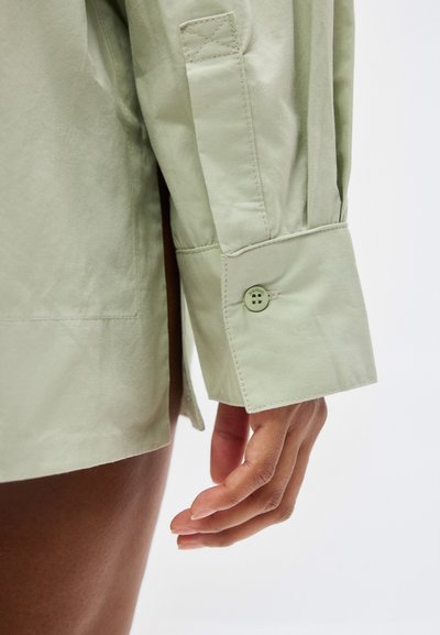 Μανσέτα πράσινου ανοιχτού πουκαμίσου με κουμπί, φορεμένη από άτομο με σκουρόχρωμο δέρμα, χέρι χαλαρό δίπλα στο μηρό σε απλό φόντο.