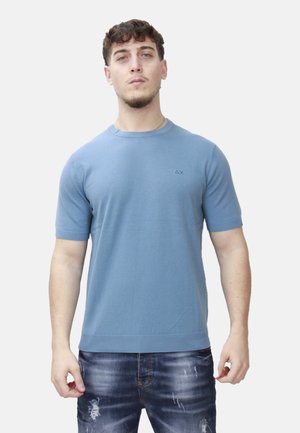 Uomo in piedi, indossa una camicia a maniche corte azzurro chiaro e jeans blu scuro consumati, volto verso l'osservatore su uno sfondo semplice.