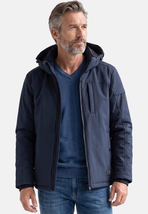 Mann mit grauen Haaren und Bart, der eine dunkelblaue Kapuzenjacke über einem blauen Pullover und Jeans trägt und leicht zur Seite schaut.