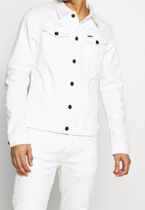 Mężczyzna ubrany w białą kurtkę jeansową z czarnymi guzikami oraz pasujące białe spodnie, stojący na jednolitym tle.