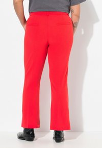 Pantalons rouges évasés avec une texture lisse et deux poches arrière. Portés avec une chemise grise et des bottes noires, en stance contre un fond blanc.