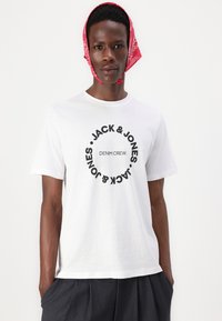 White cotton t-shirt features a circular black logo with the text "JACK & JONES" and "DENIM CREW." Model wears a red bandana.