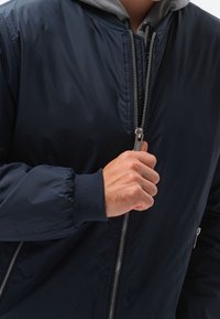 Marinblå bomberjacka med en slät, glänsande yta. Dragkedjeöppning, ribbade ärmslut och en grå huva som kiknar fram under.