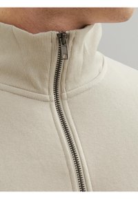 Giacca beige con zip e colletto a coste, realizzata in tessuto texturizzato. Presenta una zip metallica sul davanti. Finitura liscia, senza motivi visibili.