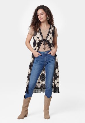 Kvinde med krøllet hår iført beige skjorte, blå jeans, brune støvler og lang sort-hvid hæklevest med blomsterprint og frynser.