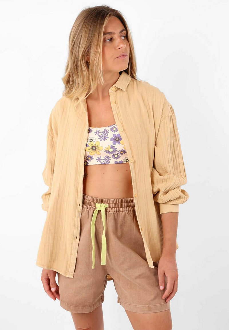 Chemise oversize beige clair avec des côtes verticales, portée sur un crop top floral et un short marron à cordon avec un lien vert lime.