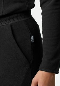 Schwarze Fleecejacke aus strukturiertem Material, mit geripptem Bund und einem kleinen Logopatch auf der Tasche. Sichtbare genähte Nähte.
