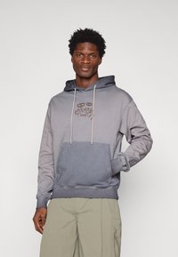 Siva dukserica s tamnoplavom kapuljačom, prednjim džepom i smešnim grafičkim logotipom u smešnoj boji. Ima sistem za zatezanjem i rebraste manžete. Mekani pamuk.