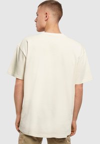 Beige oversize t-paita, jossa on lyhyet hihat, pyöreä pääntie ja sileä puuvillakangas, kuvattuna takaa vaaleanharmaata taustaa vasten.