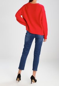 Red ribbed knit sweater with a wide fit, paired with fitted blue jeans and black stiletto heels. The sweater has a rounded neckline.