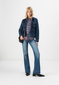 Kvinna som står i en denimjacka, rutigt skjorta, utsvängda jeans, svarta skor och axelväska mot en ren vit bakgrund.