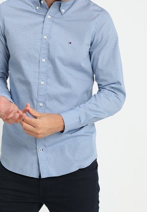 Man adjusting cuff of light blue patterned button-up shirt, wearing dark pants, against plain white background.