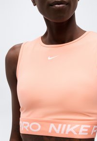 Κοντινό πλάνο ενός ατόμου που φοράει αθλητικό σουτιέν Nike Pro σε ροδακινί χρώμα με λευκό λογότυπο Swoosh στο στήθος και ελαστική ταινία με επωνυμία.