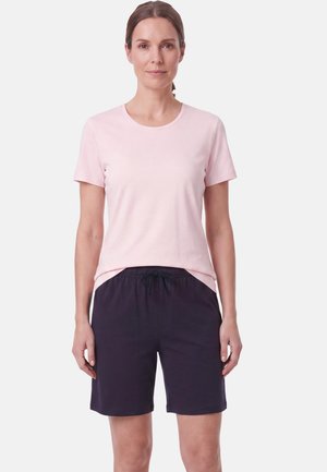 Vrouw staand met het gezicht naar voren, draagt een lichtroze T-shirt met korte mouwen en zwarte short met trekkoord tegen een effen witte achtergrond.