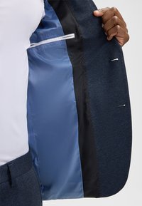 Granatowa marynarka o fakturowanej powierzchni i gładkiej, niebieskiej podszewce z satyny. Posiada kieszeń i kontrastowe przeszycia. Klasyczny, dopasowany krój.