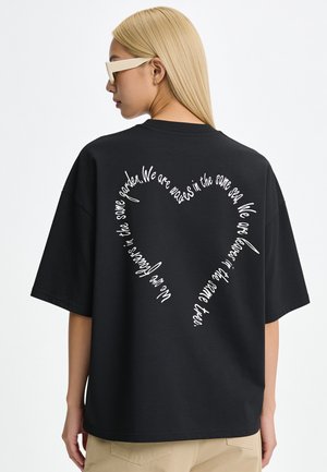 Femme aux longs cheveux blonds et lunettes de soleil, portant un t-shirt noir avec un texte blanc en forme de cœur dans le dos.