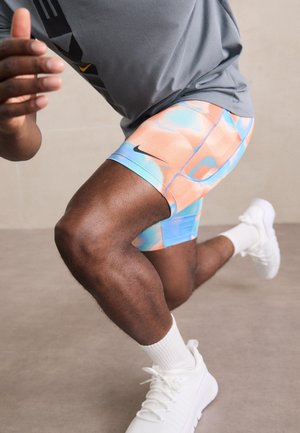 Mann in grauem Hemd und bunten Nike-Shorts, der nach vorne ausfällt, trägt weiße Socken und Turnschuhe vor einfarbigem Hintergrund.