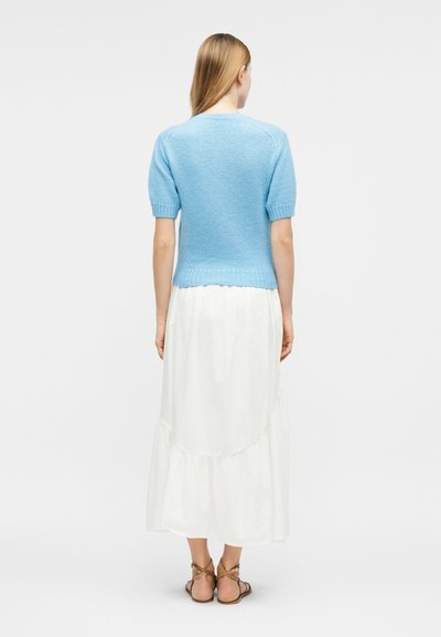 Femme debout, de dos, portant un pull à manches courtes bleu clair, une jupe blanche longueur cheville et des sandales marron à lanières.