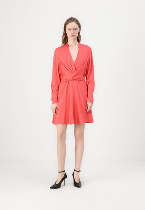 Korallenfarbiges Wickelkleid mit langen Ärmeln, tiefem V-Ausschnitt und gedrehter Detail am Bund. Kombiniert mit schwarzen Riemchen-Heels. Glatte Textur.