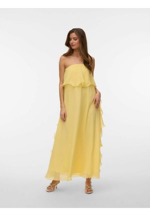 Kvinne iført en stroppeløs, flytende gul maxikjole med volangdetaljer, stående med lukkede øyne og avslappede hender langs sidene.