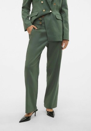Femme portant un pantalon vert olive sur mesure et un blazer assorti avec des boutons dorés, chaussures noires à talons hauts pointues, une main dans la poche.