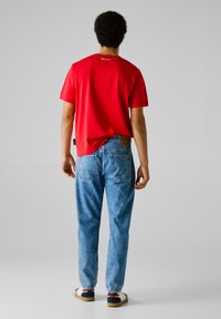 Hombre de pie de espaldas, vistiendo una camiseta roja, jeans de mezclilla claros y zapatillas blancas, contra un fondo gris liso.