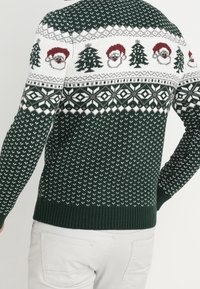 Grüner Strickpullover mit einem weißen Muster, das rote Santa-Gesichter und Tannenbäume zeigt. Gerippte Bündchen und Saum, strukturierter Stoff.