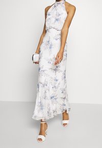 Kvinna i en vit blommig klänning med halterneck, prydd med blå och lavendelfärgade blommor, bär en vit clutch och har på sig vita högklackade sandaler.