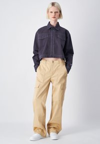 Zwarte cropped denimjas met knopen aan de voorkant en twee borstzakken, gecombineerd met beige cargobroek met zijzakken, gedragen met witte sneakers.
