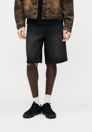 Osoba stojaca pred jednoliatym pozadim, oblecena v ciernych denimovych kratasy, bielych ponozkach, ciernych teniskach a hnedom vzorovanom sakovacom.