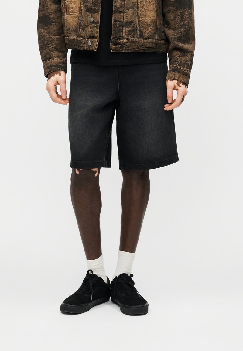 Άτομο που φοράει μαύρα τζιν σορτς, λευκές κάλτσες, μαύρα αθλητικά παπούτσια και καφέ ζακέτα με σχέδια, στέκεται μπροστά από απλό φόντο.