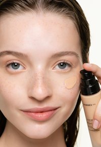 Bouteille de fond de teint dans une main, montrant une texture crémeuse tan sur la peau. Modèle avec des taches de rousseur légères, peau éclatante et maquillage naturel.