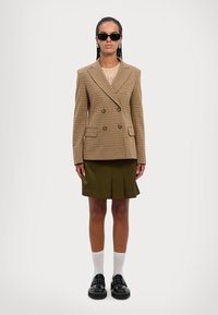 Brun houndstooth dobbeltradet jakke, kombinert med en grønn plisserte skjørt. Modellen har på seg svarte solbriller og svarte platåsko med hvite sokker.