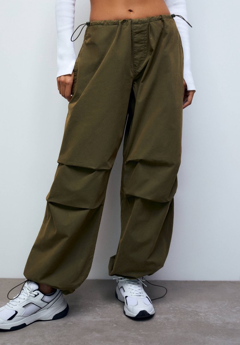 Olijfgroen, losvallende broek met een verstelbare tailleband, voorzien van twee horizontale zakken en verticale paneeldetails. Gecombineerd met witte sneakers.