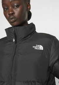 Giacca piumino nera con colletto alto, dotata di zip frontale e logo bianco ricamato nell'area del petto sinistro. Materiale liscio e leggero.