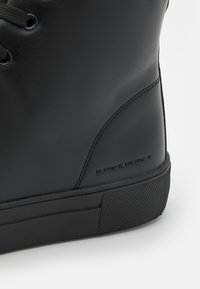 Zapatilla alta negra de cuero suave, con punta redonda, suela de goma texturizada y coordenadas geográficas en relieve en el lateral.