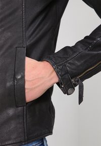 Giacca in pelle nera con design aderente, caratterizzata da una cerniera argentata, texture trapuntata sulle maniche e dettagli con cinturini regolabili.