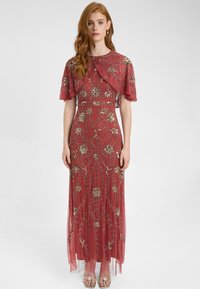 Abito lungo rosso con intricati ricami dorati, maniche a cappotto corte e una sovrapposizione trasparente. Sandali piatti e decorati completano il look.
