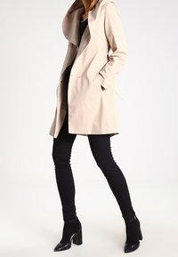 Beige sadetakki hupulla, valmistettu kevyestä kankaasta, jossa on sivutaskut ja nappisuljenta. Päällystetty mustien housujen ja kenkien kanssa.