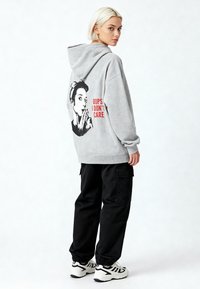 Mlada žena s kratkom plavom kosom nosi sivi hoodić s crno-bijelim grafičkim uzorkom i crvenim natpisom "OOPS I DON'T CARE", crne cargo hlače i bijele tenisice.