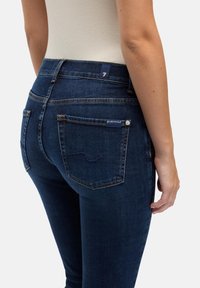 Donkere blauwe spijkerbroek met een aansluitend ontwerp, voorzien van twee achterzakken, subtiele stiksels en een merklabel aan de tailleband.