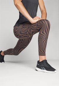 Leggings marron à motif avec un design tourbillonnant, associés à un haut de sport noir et des baskets noires. Les leggings présentent une coupe ajustée.