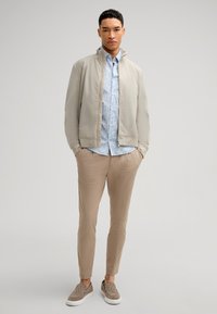 Giacca beige chiaro con zip, camicia azzurra a fantasia, pantaloni beige e scarpe slip-on texturizzate. Tessuti lisci con vestibilità casual e linee pulite.