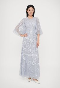 Μακρύ, ανοιχτό μπλε φόρεμα με μοτίβο λουλουδιών από παγιέτες, διάφανα μανίκια με βολάν και στρώσεις από τούλι. Διαθέτει στρογγυλή λαιμόκοψη και ρέουσα σιλουέτα.