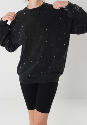 Person iført en løs sort sweatshirt med hvide prikker og sorte cykelshorts, justerer sweatshirten ved skuldrene.
