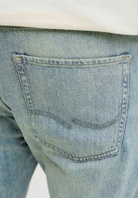 Hele sinise teksade tagatasku, millel on topeltõmblused ja peened kaardus tikandid, inimesel, kes kannab valget tükki.