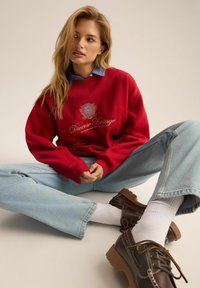 Czerwona bluza z białym haftem, jasnoniebieskie oversize'owe dżinsy i brązowe buty na sznurówki. Modelka siedzi w pozycji skrzyżowanej, zwrócona lekko w prawo.