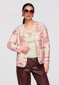 Femme portant de grandes lunettes de soleil, veste boutonnée à motif floral, haut crème drapé et pantalon en cuir marron, debout avec une main sur la veste.