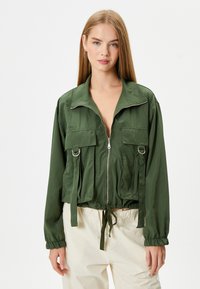 Groene cropped jack met een rits aan de voorkant, grote borstzakken en gathered manchetten. Heeft een trekkoord in de taille en glanzende hardware accenten.