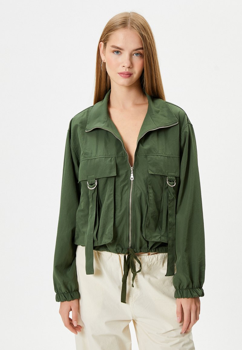 Groene cropped jack met een rits aan de voorkant, grote borstzakken en gathered manchetten. Heeft een trekkoord in de taille en glanzende hardware accenten.